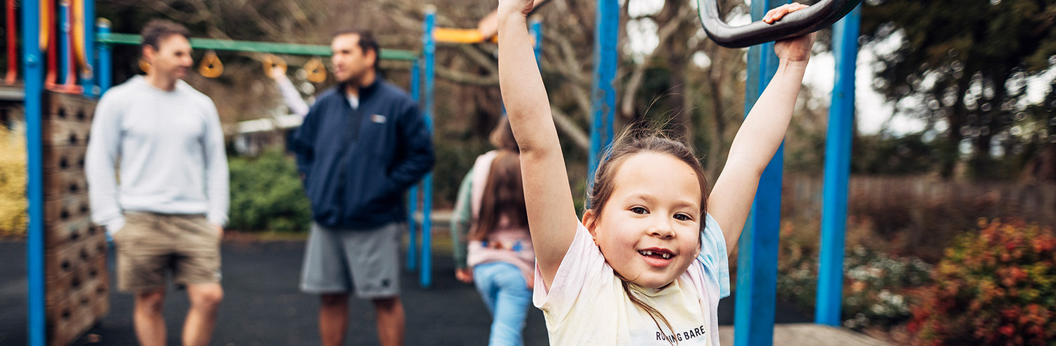 Girl smiles on playground with members of her whānau in the background.